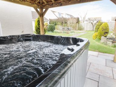 An outdoor hot tub under a wooden pergola with a garden and houses in the background at The Elms in Milford Haven