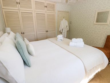 A bedroom with a bed with pillows and folded towels a robe on the door white wardrobes and a wicker chair at The Elms in Milford Haven