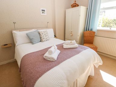 A bedroom with a double bed with pillows and folded towels a wardrobe and a wicker chair by the window at The Elms in Milford Haven