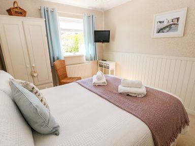 A bedroom with a bed folded towels a wicker chair a wardrobe small wall mounted television and a window with blue curtains at The Elms in Milford Haven