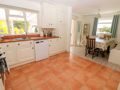 A kitchen with white cabinets dishwasher and sink next to a dining area with table chairs and a window at The Elms in Milford Haven