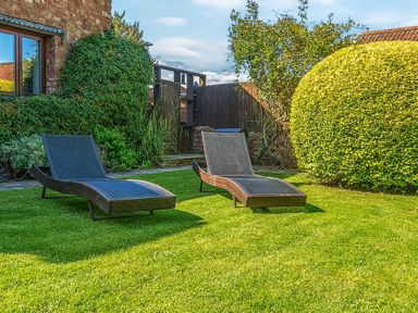 A garden with two lounge chairs on grass near a house and trimmed bushes at The Linhay in Washford near Watchet