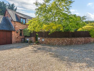 A gravel driveway with a stone building and wooden garage door next to a fenced garden at The Linhay in Washford near Watchet