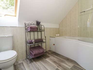 A bathroom with a toilet, a metal shelf with folded towels and lavender, and a bathtub with a glass shower screen at Squirrels Drey Little Dunham near Litcham