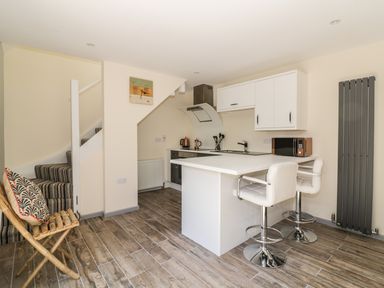 A kitchen with a white island and bar stools next to stairs with striped carpet at Squirrels Drey in Little Dunham near Litcham