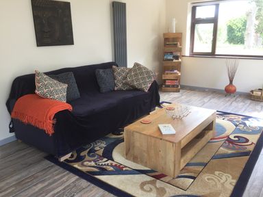 A living room with a black sofa with patterned cushions and orange throw a wooden coffee table on a patterned rug a wooden bookshelf and a window at Squirrels Drey in Little Dunham near Litcham