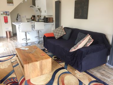 A living room with a sofa covered with a dark cloth and patterned cushions a wooden coffee table two white bar stools and a kitchen counter at Squirrels Drey Little Dunham near Litcham