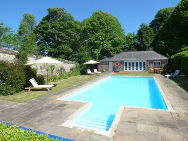 An outdoor swimming pool with lounge chairs and umbrellas surrounded by trees and a stone building at Parlour Cottage in Marazion