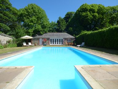 An outdoor swimming pool with lounge chairs and an umbrella near a small stone building with trees behind at Parlour Cottage in Marazion