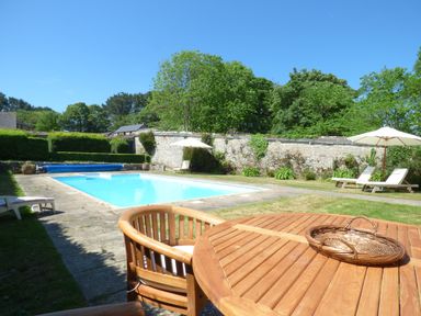 A backyard with a swimming pool surrounded by stone pavement chairs with umbrellas and a wooden table with a basket at Parlour Cottage in Marazion