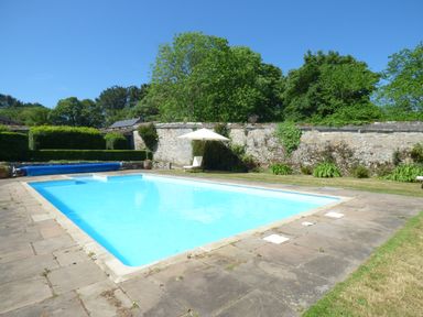An outdoor swimming pool surrounded by stone paving and grass with a stone wall and trees at Parlour Cottage in Marazion