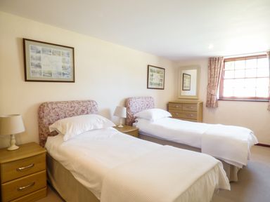 A bedroom with two single beds bedside tables lamps chest of drawers a window and framed pictures at Parlour Cottage in Marazion