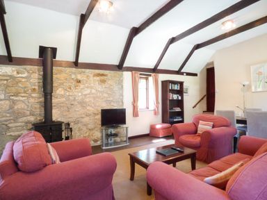 A living room with red armchairs, a wooden coffee table, a wall-mounted stove, a stone feature wall, a television, a bookshelf, and a dining area at Parlour Cottage in Marazion