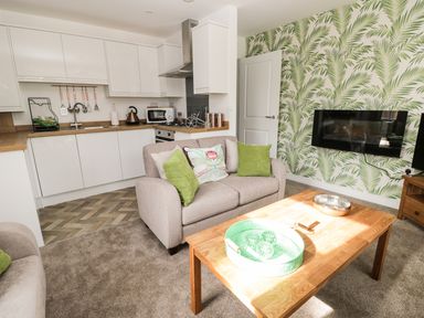 A living room and kitchen with a beige sofa, green cushions, a wooden coffee table, and a palm leaf wallpaper at Manor Heath Apartment 1 in Scarborough