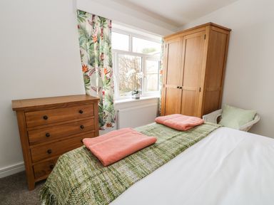 A bedroom with a wooden chest of drawers a wooden wardrobe a bed with green and coral bedding and a window with leaf patterned curtains at Manor Heath Apartment 1 in Scarborough