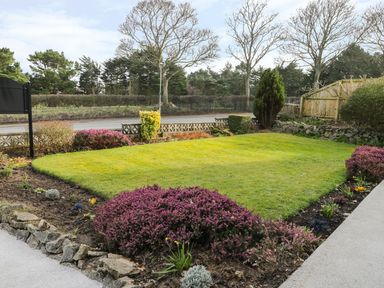 A garden with green grass surrounded by flower beds and shrubs with trees and a road in the background at Manor Heath Apartment 1 in Scarborough