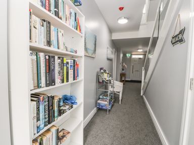 A hallway with a white bookshelf filled with books and a small metal rack with magazines and board games at Manor Heath Apartment 4 Scarborough