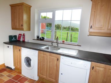 A kitchen with wooden cabinets washing machine dishwasher and a window showing a grassy field at The Slate House in Cahersiveen County Kerry