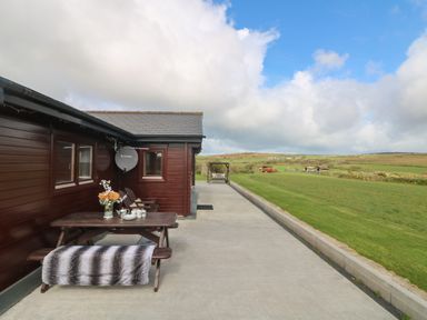 An outdoor patio area with a wooden picnic table with a flower vase and cups and a bench with a blanket beside a brown wooden building with fields and blue sky in the background at Chywolow Lodge in Penzance