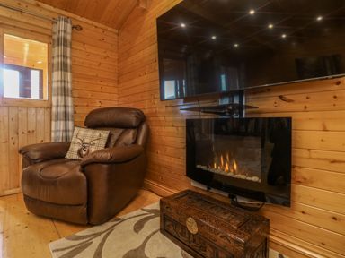 A wooden room with a brown leather armchair a plaid cushion a wall-mounted electric fireplace a wooden chest and a mounted television at Chywolow Lodge in Penzance