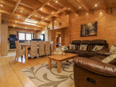A living area with a dining table and chairs next to a kitchen and a leather sofa with cushions in a wooden cabin at Chywolow Lodge in Penzance