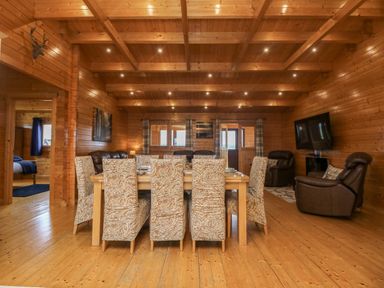 A dining area with eight patterned chairs around a wooden table and a living area with leather chairs and a wall mounted TV in a wooden cabin at Chywolow Lodge in Penzance