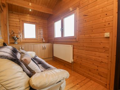 A wooden room with a cream sofa covered by a blanket and pillows next to windows and a radiator at Chywolow Lodge in Penzance