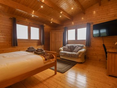 A bedroom with a wooden bed, sofa, wardrobe, TV, and windows with curtains at Chywolow Lodge in Penzance