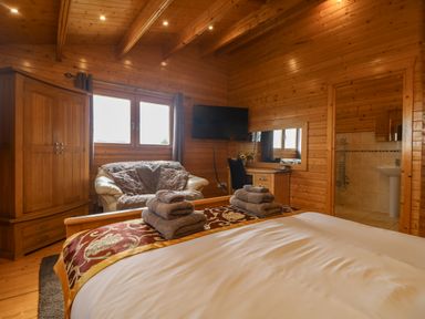 A bedroom with a bed with folded towels a sofa a wooden wardrobe a desk with a chair and a wall-mounted TV at Chywolow Lodge in Penzance