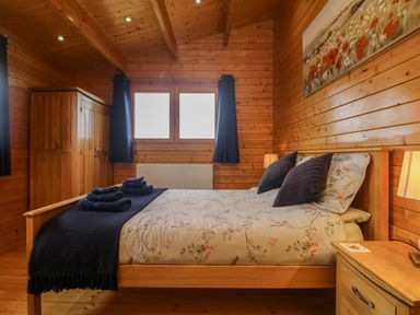 A bedroom with a wooden bed and side table two windows with curtains and a wooden wardrobe at Chywolow Lodge in Penzance