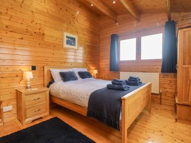 A wooden bedroom with a bed towels on the bed bedside table lamps window with curtains and a wardrobe at Chywolow Lodge in Penzance
