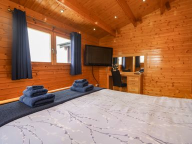 A bedroom with a bed towels on it a wall mounted tv a dressing table and wooden walls at Chywolow Lodge in Penzance