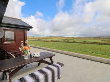 An outdoor wooden picnic table with flowers cups and plates next to a red wooden building overlooking green fields at Chywolow Lodge in Penzance