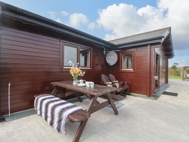 An outdoor wooden picnic table with a blanket and flowers next to a dark wooden building at Chywolow Lodge in Penzance