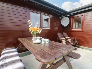 An outdoor patio area with a wooden picnic table set with mugs plates a teapot and a vase of flowers against a wooden wall with two chairs and a satellite dish at Chywolow Lodge in Penzance