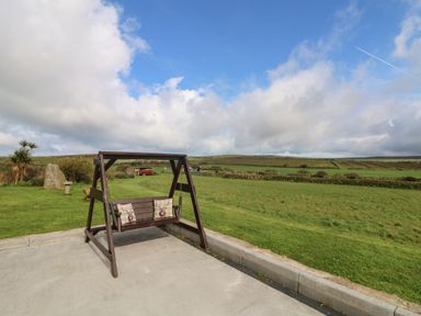 A wooden swing with pillows on a concrete surface with a grassy field and cloudy sky in the background at Chywolow Lodge in Penzance