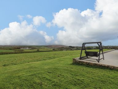 A swing on a patio overlooking a grassy field with distant hills and clouds at Chywolow Lodge in Penzance