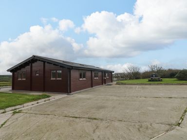 An exterior view of a brown wooden building with multiple windows a concrete driveway and a grassy area with a picnic table at Chywolow Lodge in Penzance