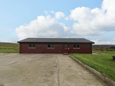 A single story building with four windows and one door surrounded by a concrete driveway and grass at Chywolow Lodge in Penzance