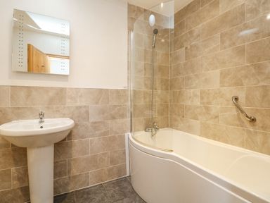 A bathroom with a white sink and mirror and a bathtub with curved glass shower screen and beige tiles at Mill in Nancegollan nr Porthleven