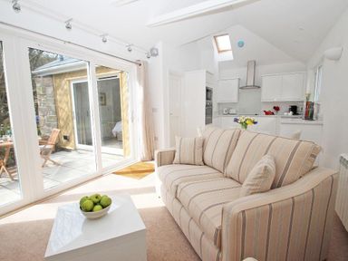 A living area with a striped sofa and bowl of green apples on a table next to a kitchen with white cabinets at Cot Valley Cottage in St Just