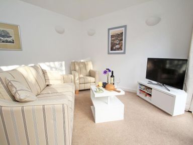 A living room with striped sofa and armchair white coffee table with food and wine and a television on a white stand at Cot Valley Cottage in St Just