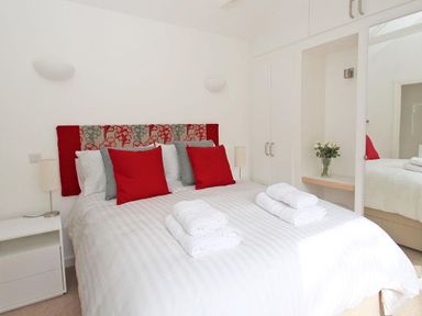 A bedroom with a double bed with white bed linen and red and grey pillows bedside tables and lamps and built in wardrobes with a mirror and flowers in a vase at Cot Valley Cottage in St Just