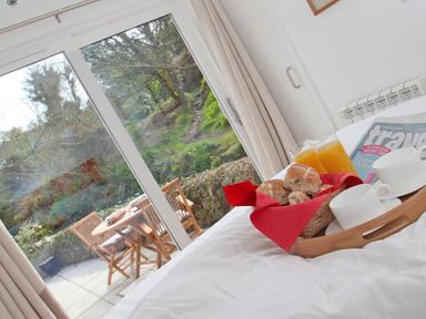 A breakfast tray with rolls orange juice and cups on a bed next to a glass door with outdoor table and chairs visible beyond Cot Valley Cottage St Just
