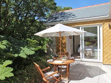 An outdoor patio with a wooden table and chairs under a white umbrella next to a sliding glass door surrounded by plants at Cot Valley Cottage in St Just