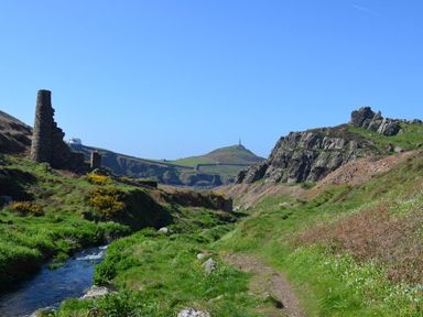 A grassy valley with a stream and stone ruins with hills and a hilltop monument in the distance at Cot Valley Cottage in St Just