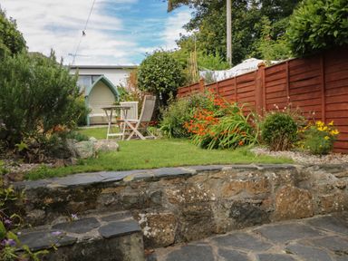 A garden with a stone patio green grass wooden chairs a table and a red fence at Pippin in Lelant