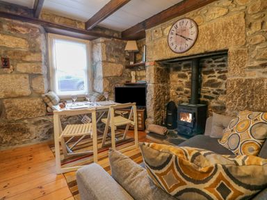 A room with a dining table and chairs next to a window a stone fireplace with a wood stove a wall clock above the fireplace and a sofa with patterned cushions at Pippin in Lelant