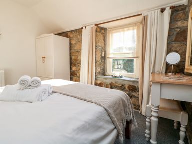 A bedroom with a bed covered in white linens and towels a white wardrobe a stone wall window with curtains and a small desk with a mirror at Pippin in Lelant
