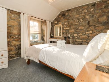 A bedroom with a bed with white linens and rolled towels on it stone walls a window with curtains and a wooden dresser with a mirror at Pippin in Lelant